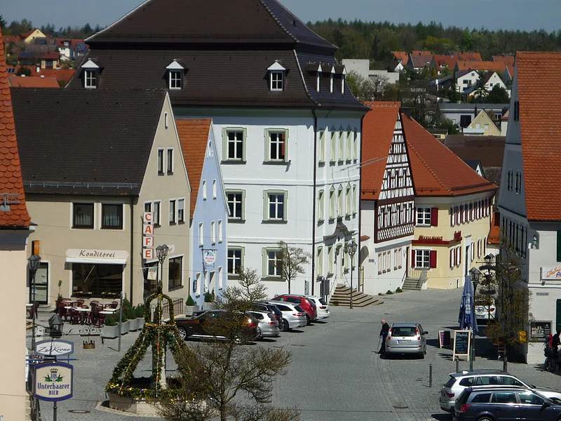 Café Konditorei Wenninger - im Herzen der Stadt Monheim Ansicht einer gepflasterten Innenstadt mit historischen Gebäuden, geparkten Autos und einem geschmückten Oster-Brunnen. Im Hintergrund sind Wohnhäuser zu sehen.