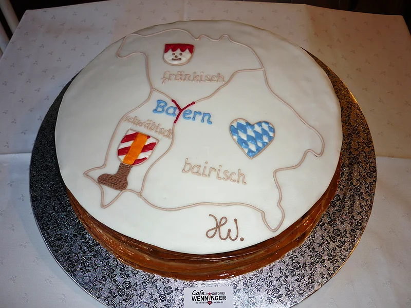 "Drei-Länder-Eck-Torte" Café Wenninger Runde Torte auf einer silbernen Unterlage seht auf einem Tisch mit weißem Tischtuch. Auf der Torte weiße Glasur und eine Karte von Bayern mit Dialektbezeichnungen und den dazugehörigen Symbolen darauf.