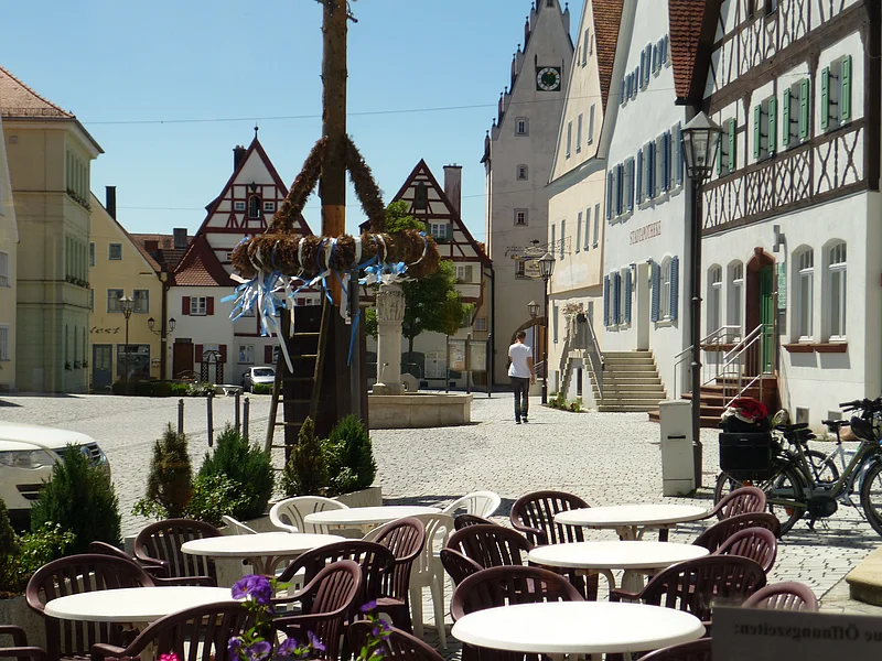 Café Konditorei Wenninger - das Café am Marktplatz Ein Marktplatz mit Tischen und Stühlen eines Cafés im Vordergrund, Fachwerkhäusern im Hintergrund und einem Maibaum in der Mitte des Marktplatzes. Eine Person läuft auf dem Markplatz.