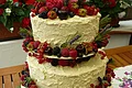 Hochzeitstorte "Frucht" Café Wenninger Dreistöckige Torte mit weißer Creme, dekoriert mit Beeren, Kirschen, Lavendel und Rosmarinzweigen steht auf einem Tisch. Im Hintergrund ein Garten mit bepflanzten Blumentöpfen und grüner Wiese.