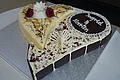 "Hochzeitsherz" Café Wenninger Herzförmige Hochzeitstorte mit weißer und dunkler Schokolade, beschriftet mit „Ingrid & Martin“ und dekoriert mit zwei roten Rosen und verschiedener Schokoladen-Dekoration, steht auf einer silbernen Unterlage auf einem Tisch.