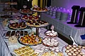 "Süßes Buffet" Café Wenninger Buffet mit verschiedenen süßen Kuchen, Gebäck und Obsttörtchen auf mehreren Etageren und Tellern auf einem Tisch aufgedeckt. Im Hintergrund sind aufgestapelte beleuchtete Teller und Tassen mit zwei Kaffeekochern.