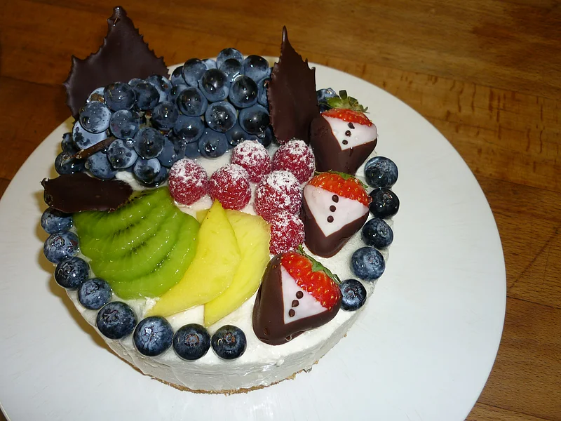 "Tutti Frutti Torte" Café Wenninger Obsttorte mit weißer Creme, obendrauf Trauben, gezuckerten Himbeeren, Kiwi, Ananas, Blaubeeren und Erdbeeren in Schokoladenmantel auf einem weißem Teller serviert.