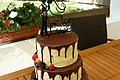 "Naked Cake Frucht" Café Wenninger Vierstöckige Hochzeitstorte mit runterlaufender Schokoladenglasur und weißer Creme, verschiedenen Beeren auf drei Stockwerken und einem Brautpaar-Topper mit Schriftzug „Mr & Mrs“ oben drauf. Im Hintergrund ein Garten mit bepflanzten Blumentöpfen und grüner Wiese.