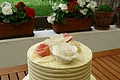 Hochzeitstorte Café Wenninger Zweistufige weiße Hochzeitstorte mit Rosen aus Zuckerguss verziert steht auf einem Holztisch im Garten. Im Hintergrund bepflanzte Blumentöpfe und grüne Wiese.