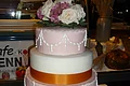 Hochzeitstorte "Klassisch" Café Wenninger Vierstöckige Hochzeitstorte mit weißer und rosa Glasur, orangene-gelbem Band um zwei Stockwerke und einem rosa-weißen Blumenarrangement obenauf.