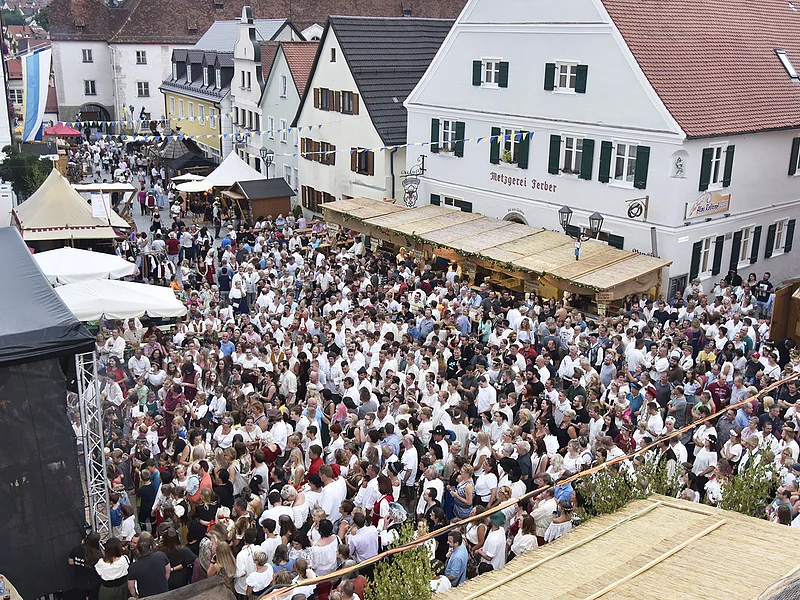 Große Menschenmenge auf dem historischen Stadtfest in der Altstadt von Monheim, mit Ständen und historischen Gebäuden im Hintergrund. Eine blau/weiße Flagge hängt an einem Gebäude.