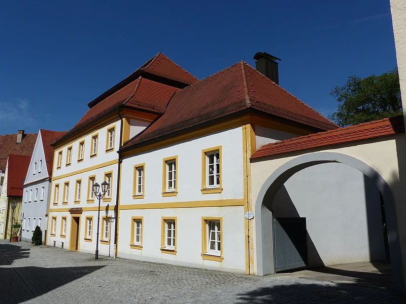 Pfarr- und Stadtbücherei Monheim Historisches weißes Gebäude, die Pfarr- und Stadtbücherei in Monheim, mit gelben Fensterrahmen und rotem Dach, daneben ein Torbogen auf gepflasterter Straße. Links vom Gebäude weitere Häuser in blau und grün.