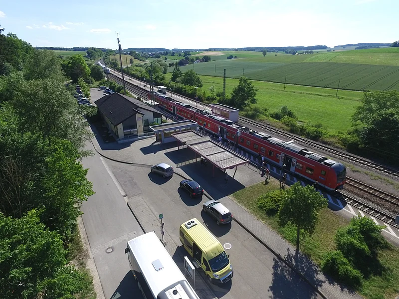 Luftaufnahme eines Bahnhofs mit rotem Zug, umgeben von Feldern und Bäumen. Stehende Autos und Busse sind zu sehen.