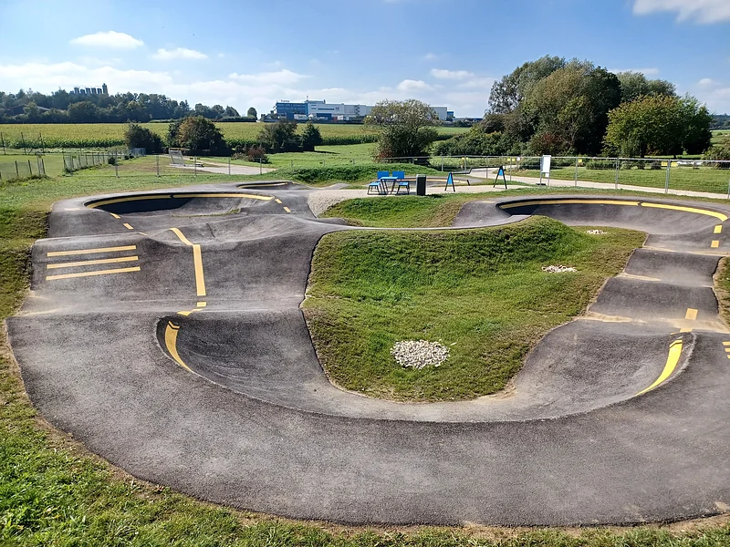 Pumptrack Monheim Asphaltierter Pumptrack mit gelben Markierungen in der Mitte, umgeben von grüner Landschaft und Bäumen im Hintergrund.