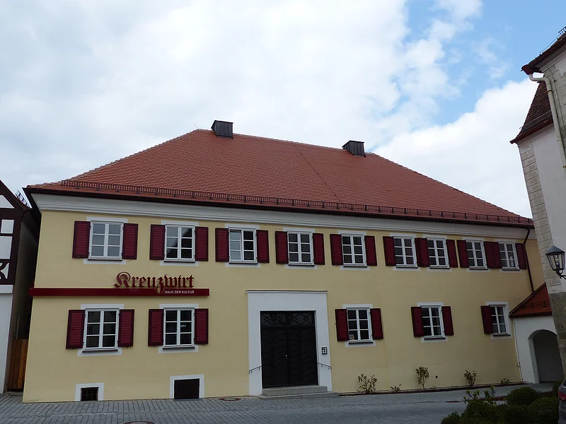 Kreuzwirt "Haus der Kultur" Monheim Gelbes zweistöckiges Gebäude mit vielen Fenstern mit jeweils zwei roten Fensterläden an jedem Fenster, einem rotem Ziegeldach und dem Schriftzug „Kreuzwirt - Haus der Kultur“ links vom Eingang. In der Mitte eine große braune zweiflüglige Holztür. Vor dem Gebäude eine Straße, im Hintergrund blauer Himmel mit Wolken.