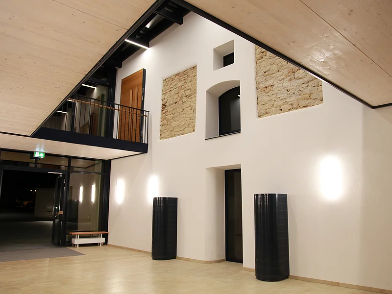 Kreuzwirt "Haus der Kultur" Monheim Heller Innenraum mit heller verkleideter Holzdecke, weißen Wänden und hellem Bodenbelag. Zwei schwarze Säulen rechts an der Wand und offener Glastür bei Nacht. Oben im offenem Obergeschoss zwei Teilbereiche einer freigelegten Steinmauer in der Wand, eine braue Holztür und schwarzes Metallgeländer.