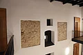 Kreuzwirt "Haus der Kultur" Monheim Innenraum mit weißer Wand, darin zwei Ausschnitte einer freigelegten Steinmauern und mehreren hellen Türen aus Holz mit schwarzen Metallgeländern. Oben an der Decke dunkle Holzbalken zu sehen.