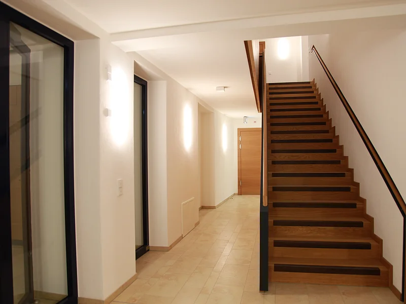 Kreuzwirt "Haus der Kultur" Monheim Flur mit beleuchteten weißen Wänden durch Wandleuchten links im Bild und einer Glastür. Eine Holztreppe, die nach oben führt rechts im Bild. Hinten im Gang eine helle Holztür, heller Bodenbelag aus Fließen.