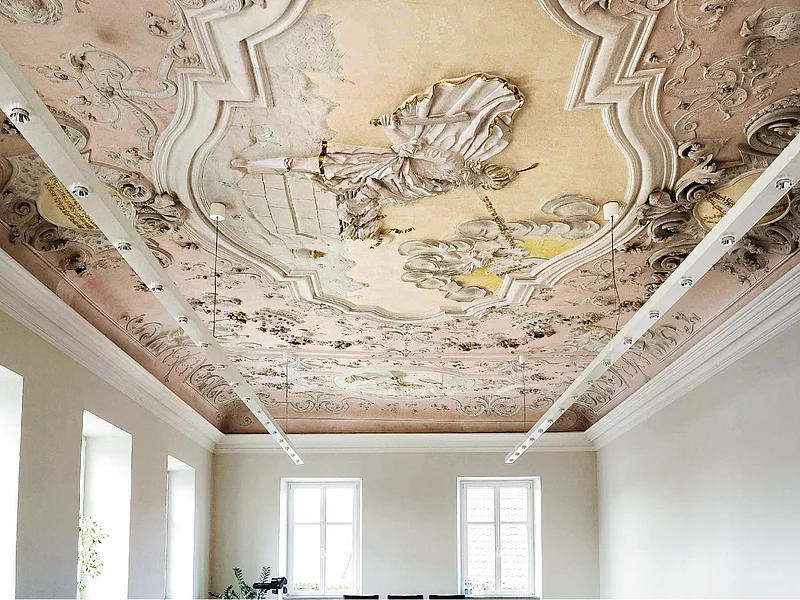 Farbig gestaltete Decke mit barocker Stuck- und Freskoverzierung in einem hellen Raum mit weißen Wände, vielen Fenstern und moderner an der Decke hängender Beleuchtung.