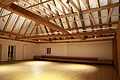 Kreuzwirt "Haus der Kultur" Monheim Beleuchteter leerer Raum mit offener Holzbalkendecke, hellem Holzboden und weißen Wänden, Fenster, Tür und eine Heizung an der Wand auf der linken Seite.