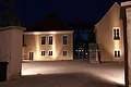 Kreuzwirt "Haus der Kultur" Monheim Beleuchteter Innenhof mit zwei gelben Gebäuden, einer schwarzen Metall-Treppe und einer hohen weißen Mauer und gepflasterten Hof bei Nacht. Dunkler Himmel im Hintergrund.