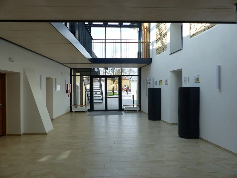 Kreuzwirt "Haus der Kultur" Monheim Heller Eingangsbereich eines großen Raums mit Ausgang nach hinten ins Freie, Ausgangstür bestehend aus Glas. Zwei schwarze Säulen an der Wand rechts im Bild. Zwei Heizkörpern mit einer Sitzauflage darauf und einem offenem Obergeschoss mit schwarzem Metallgeländer darüber. Im Freien links im Bild eine schwarze Treppe führend in das Obergeschoss zu sehen.
