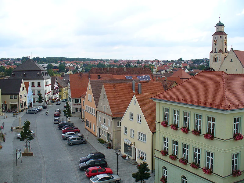 Blick auf eine Straße in der Monheimer Innenstadt mit geparkten Autos an der Straße entlang und Fachwerkhäusern mit roten Dächern bei bewölktem Himmel. An der Straße einzelne Bäume gepflanzt, ein Brunnen auf der linken Seite ist zu sehen und auf der rechten Seite eine Kirche.