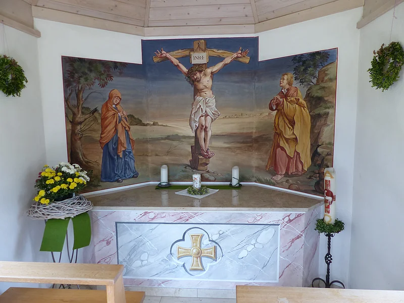 Altar in einer Kapelle mit Kreuzigungsszene vom Jesus am Kreuz, Maria links, Johannes rechts an einer weißen Wand. Drei Kerzen stehen auf dem Altar und eine große Kerze rechts davon auf einem Kerzenständer. Blumenarrangement links am Altar und zwei grüne Kränze an der Wand angebracht.