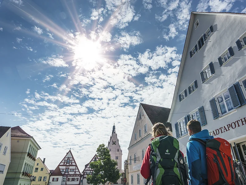 Wanderer in Monheim Zwei Personen mit Rucksäcken gehen durch eine Stadt mit Fachwerkhäusern unter einem sonnigen Himmel. Auf dem Haus im Vordergrund steht die Aufschrift "Stadtapotheke".