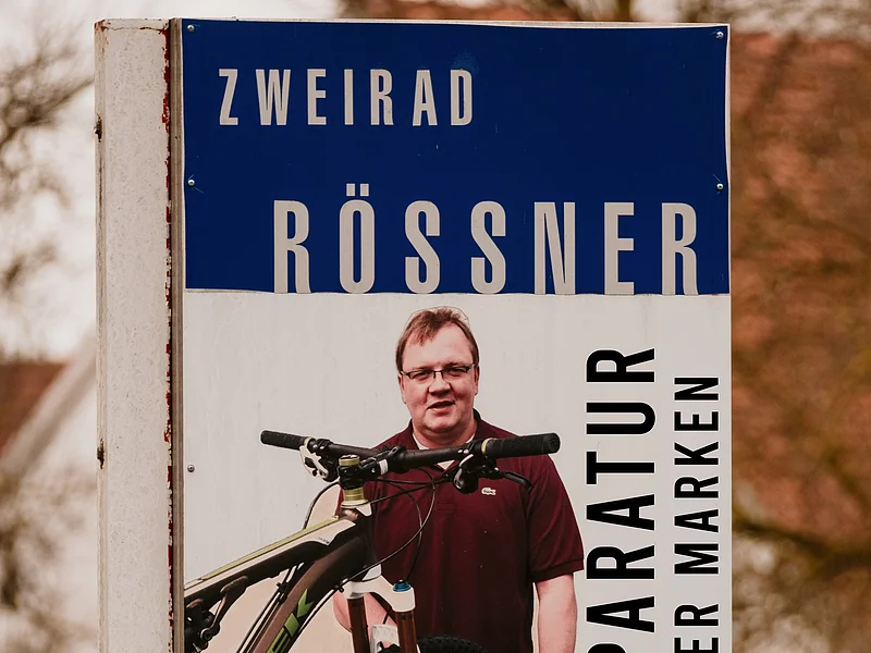 Zweirad Rößner - Reparaturservice für Fahrräder aller Marken Werbeschild für Fahrradreparatur mit Mann und Fahrrad, Text „Zweirad Rössner Reparatur aller Marken“ und Telefonnummer.