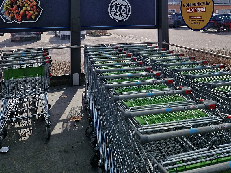 Einkaufswägen am Aldi-Süd Überdachter Einkaufswagenbereich aus Glas mit Aldi-Werbung darin und geparkten Autos im Hintergrund.
