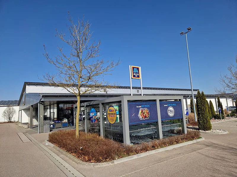 Aldi Süd - Außenansicht der Filiale in Monheim Außenansicht eines Aldi Süd Supermarkts mit Einkaufswagenunterstand, großem umgebenden Parkplatz und kahlem Bäumen bei klarem Himmel.