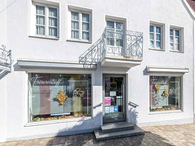 Fassade eines Friseursalons mit Fenstern, zwei Schaufenstern und einem Eingang. Am grau-blauem Gebäude die Aufschrift "Frisurentreff" und oberhalb der Eingangstür ein kleiner Balkon. Vor dem Gebäude ein gepflasterter Fußgängerweg.