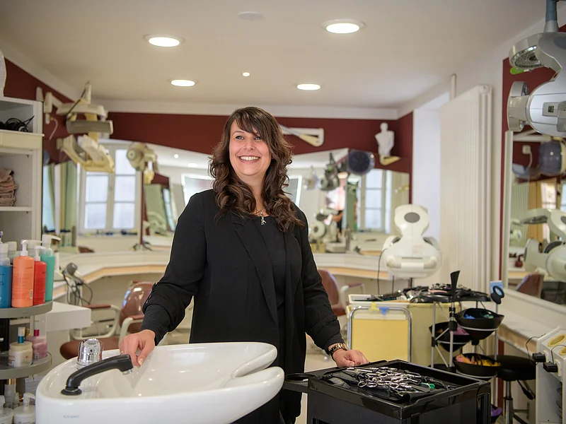 Frau steht lächelnd in einem Friseursalon neben einem Waschbecken und einem Wagen mit Friseurzubehör.