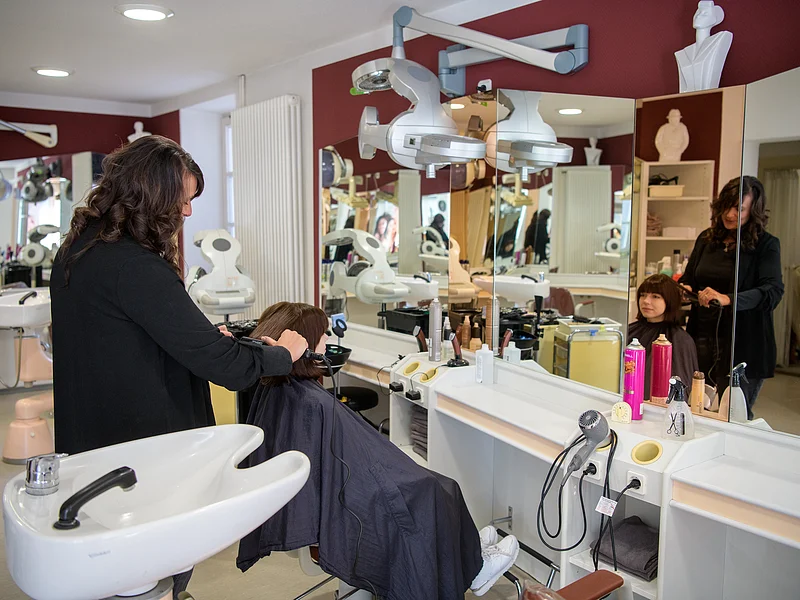 Friseurin stylt Haare einer dunkelhaarigen Kundin vor großem Spiegel in einem Friseursalon mit Waschbecken und Haartrockner.