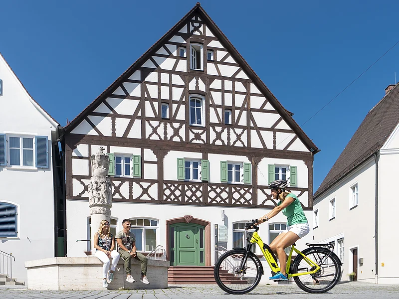 Der Marktplatz im Herzen der Stadt Monheim Der Marktplatz in Monheim: ein Fachwerkhaus zwischen zwei Gebäuden, ein Fahrradfahrer im Vordergrund fährt auf dem gepflastertem Platz, zwei Personen sitzen auf dem Rand eines Brunnens vor dem Fachwerkhaus. Auf dem linken Gebäude steht die Aufschrift "Stadtapotheke", auf dem rechten Gebäude die Aufschrift "Sparkasse".