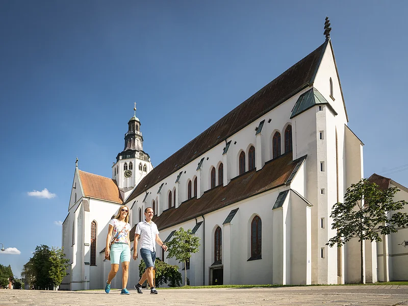 Marienmünster in Kaisheim Zwei Personen gehen vor einer großen weißen Kirche mit Turm und blauem Himmel spazieren.
