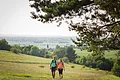 Wandern bei Huisheim Zwei Wanderer mit Rucksäcken gehen auf einem Weg durch eine grüne Wiesenlandschaft bei klarem Himmel.