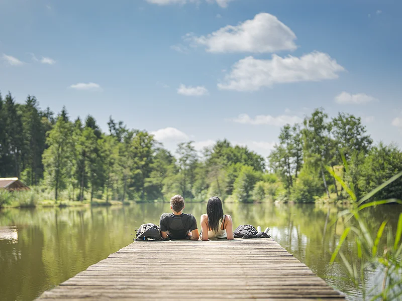 Monheimer Alb - Natur erleben Zwei Personen sitzen auf einem Steg an einem See, umgeben von Bäumen und unter blauem Himmel. Im Hintergrund links im Bild eine kleine Holzhütte zu sehen.