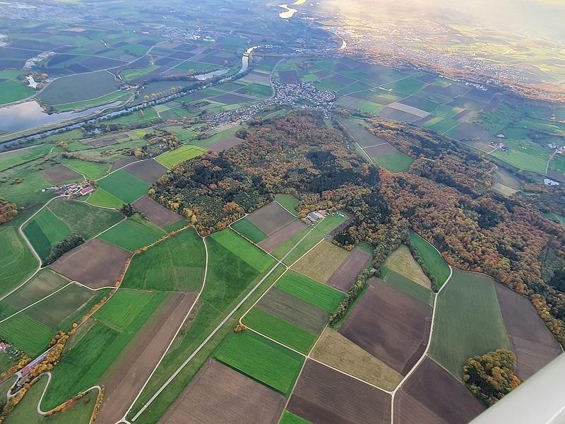 Luftaufnahme von Feldern, Waldflächen und einem Fluss bei Sonnenlicht, mit Dörfern und Straßen.