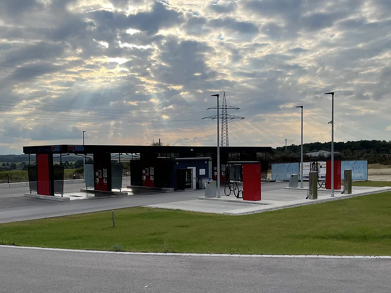 Freie Autowaschanlage mit mehreren Waschplätzen unter bewölktem Himmel und Sonnenstrahlen. Davor eine grüne Rasenfläche.