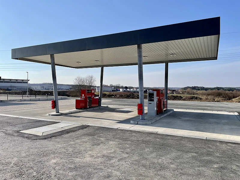 Leere Tankstelle mit zwei roten Zapfsäulen auf einem gepflasterten Platz, unter einem großen Dach bei klarem Himmel.