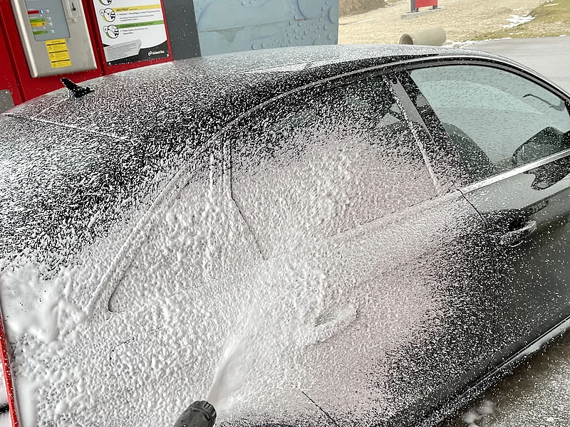 Schwarzes Auto wird an einer Waschstation mit Schaum aus einer Hochdruckpistole gereinigt.