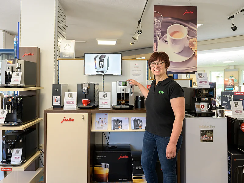 Elektro Gerstner - Elektrogeräte Frau steht in Elektronikgeschäft vor Jura-Kaffeemaschinen und zeigt auf eine Maschine mit Kaffeeauslauf.
