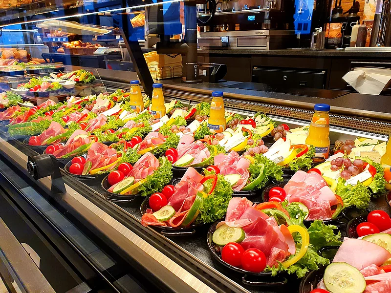Auslage mit verschiedenen belegten Tellern, gefüllt mit Aufschnitt, Käse, Schicken, Wurst, Gemüse und Orangensaftflaschen. Im Hintergrund Kaffeemaschinen zu sehen.
