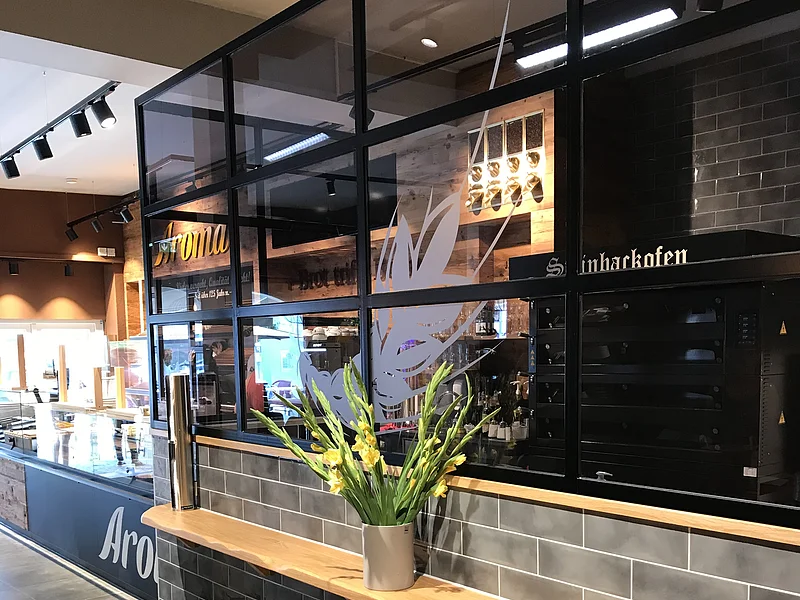 Innenraum eines modernen Cafés mit grauen Fliesen, einer Holzablage und einer Vase darauf mit gelben Blumen. Im Hintergrund ist eine Verkaufstheke und Küchengeräte.