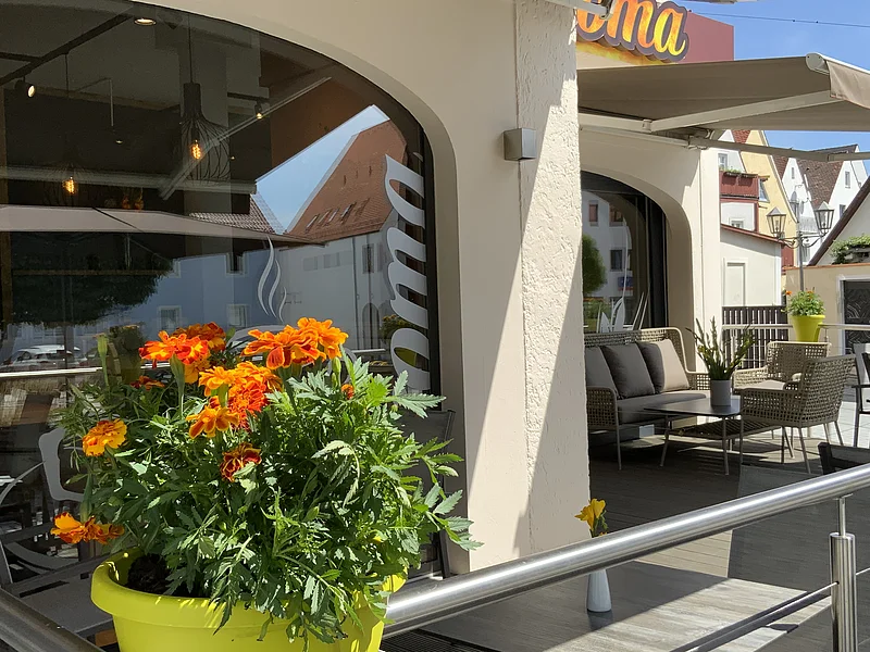 Blumentopf mit orangefarbenen Blumen auf dem Geländer einer Terrasse vor einem Café mit grauen Sitzgelegenheiten und Sonnenschirm. Auf der Fensterscheibe steht der Text "Aroma".