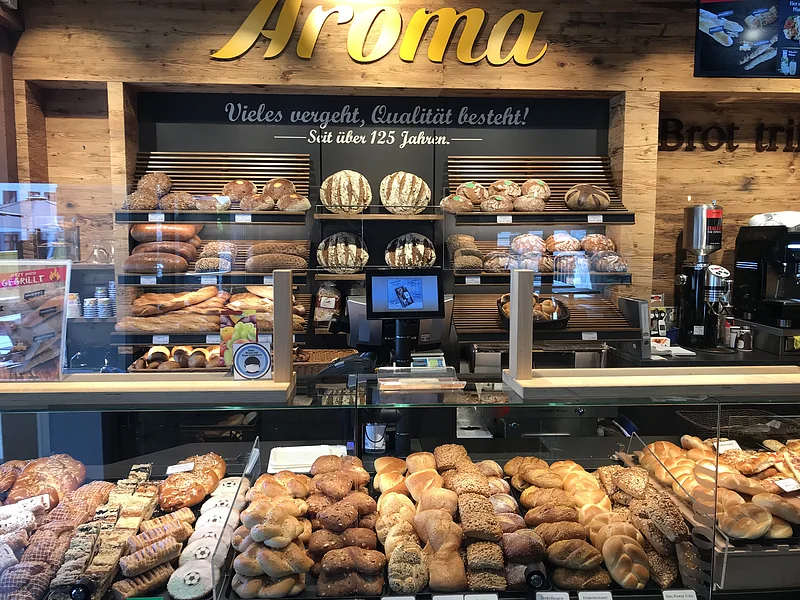 Bäckerei-Theke mit einer Vielzahl von Broten und Backwaren hinten auf Regalen und in Auslagen. An der Wand gelbe Aufschrift "Aroma".