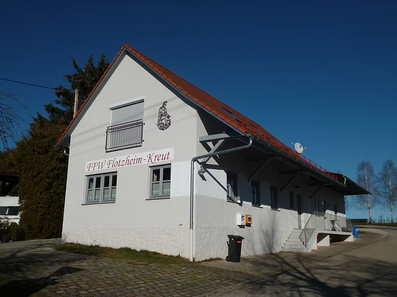 Feuerwehrhaus mit Aufschrift „FFW Flotzheim-Kreut“ und rotem Ziegeldach bei klarem Himmel. Umgeben von Bäumen, einem gepflastertem Platz und blauem Himmel.