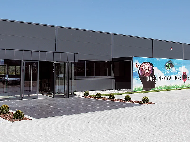 beko Group - das Schulungszentrum "beko box" in Monheim Eingang eines modernen grauen Gebäudes mit Glasfront, Flachdach und Werbebannern an der Fassade, bei strahlend blauem Himmel. Vor dem Eingang zwei bepflanzte Beete links und rechts, umgeben vom großen gepflastertem Hof.