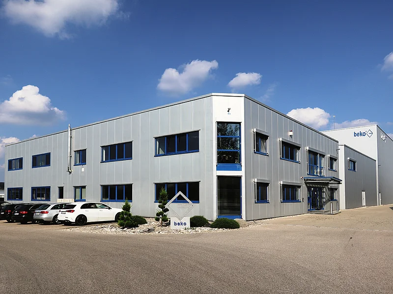 beko GmbH Gebäude Gebäude mit silberner Fassade und blauen Fenstern, davor geparkte Autos und ein Schild mit der Aufschrift „beko“ vor dem Gebäude. Im Vordergrund gepflasterter Hof und blauer Himmel im Hintergrund.