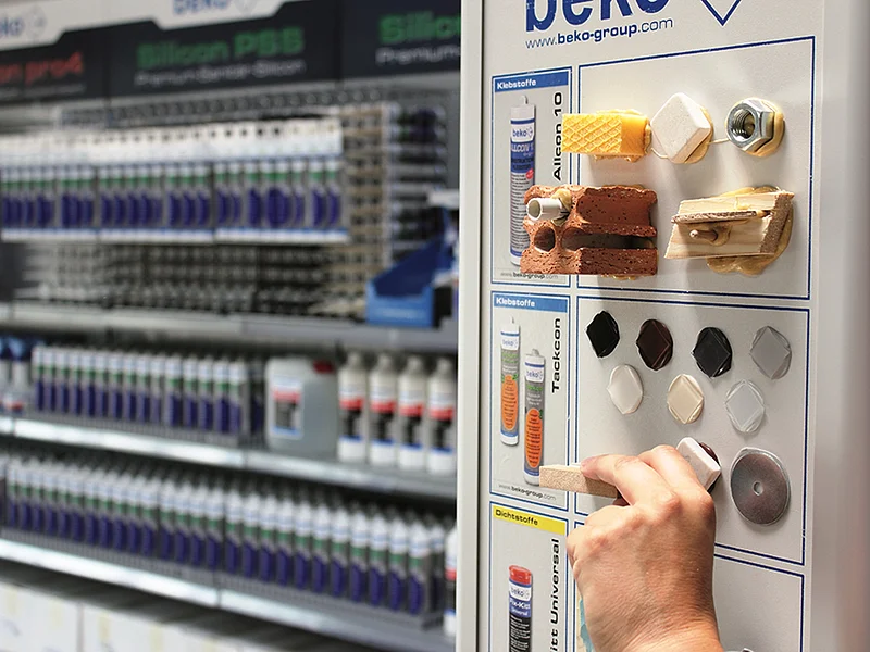 beko GmbH Verkauf Hand hält Musterstück an ein Display mit Klebstoffproben und ausgestellten Materialien in Regalen eines Fachgeschäfts.
