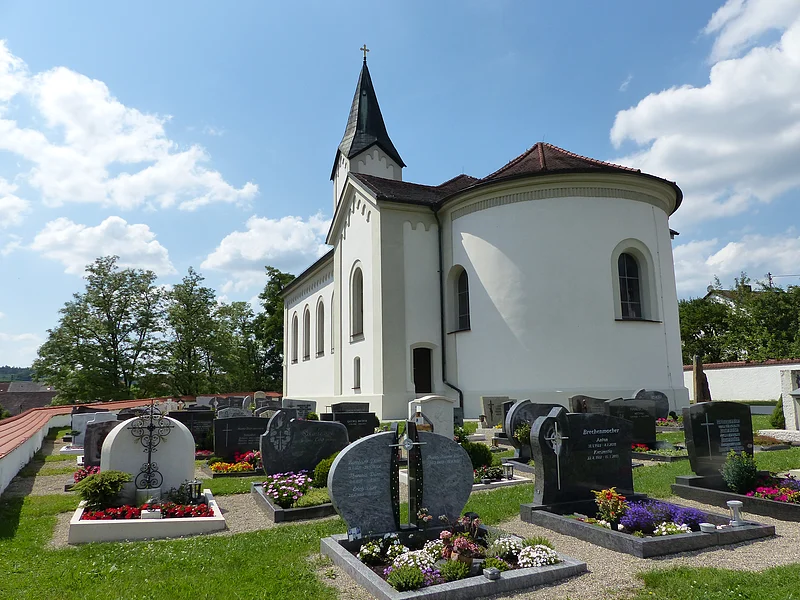 Weiße Pfarrkirche mit abgerundetem Gebäude, dunklem Dach und einem kleinen Turm mit schwarzem Dach. Anliegend ein Friedhof  mit Grabbepflanzung und Grabsteinen bei sonnigem Himmel und wenigen Wolken.