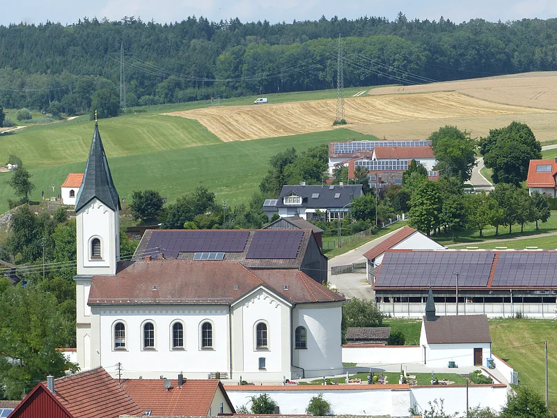 Pfarrkirche mit abgerundetem Gebäude und einem Turm, Felder und Wald im Hintergrund an einem sonnigen Tag. Umliegende Häuser mit Solarpaneelen.